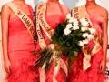 Tres mujeres vestidas de gala posan con coronas y fajas en una competencia de belleza. La imagen muestra detalles como vestidos rojos, coronas y fajas con nombres de eventos.