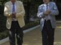 Dos hombres mayores caminan por un camino de piedra, uno con chaqueta beige y corbata azul, el otro con chaqueta de rayas grises y corbata beige. Ambos llevan pantalones oscuros y zapatos de vestir. El entorno es verde con árboles en el fondo, sugiriendo un ambiente tranquilo y sereno.