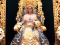 Imagen de una figura religiosa vestida con un manto azul y dorado, adornada con joyas y una corona. La figura lleva un manto azul con detalles dorados, una saya bordada y un cinturón. La figura está sentada en un trono dorado con flores blancas a los lados.