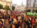 Una multitud de personas reunidas en una calle, con algunos portando sombrillas y otros mirando hacia adelante. En el fondo se pueden ver edificios históricos con detalles arquitectónicos.