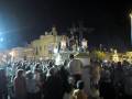 Fotografía nocturna de una procesión religiosa en un pueblo con edificios antiguos. La imagen muestra a una multitud de personas participando en la procesión, con un altar y una estatua iluminada como el centro de atención. La escena se desarrolla en un entorno urbano con edificios y luces de fondo.