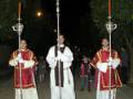 Tres sacerdotes vestidos con hábitos blancos y rojos, portando cruces en sus manos, caminan por una calle nocturna. La escena transmite solemnidad y rituales religiosos.