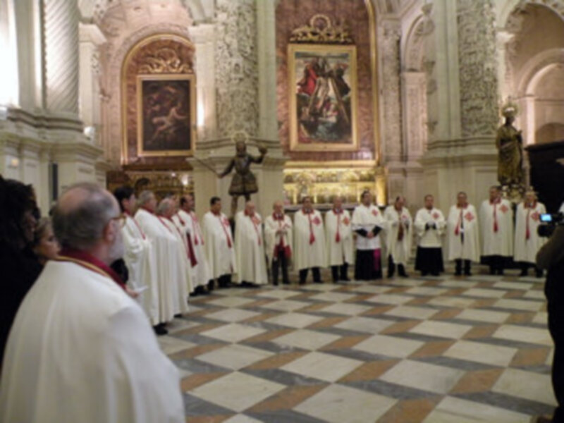 Imagen de un grupo de sacerdotes vestidos con hábitos blancos y rojos, posiblemente durante una ceremonia religiosa en un edificio histórico con arcos y vitrales.