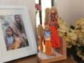 Una figura de muñecas de arcilla está hecha con un vestido rojo y una falda naranja, junto a otra figura de muñecas con un vestido azul y una falda naranja. En el fondo, hay flores amarillas y un marco de fotos con una imagen de dos personas.