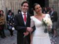 A una pareja casada en un evento formal, posando frente a una iglesia. La novia lleva un vestido blanco con un ramo de flores y el novio, una chaqueta negra con corbata roja. La escena está rodeada por otros invitados vestidos formalmente, algunos con sombreros.