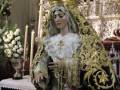 A una figura vestida con trajes tradicionales y ornamentos elaborados, posiblemente en un evento religioso o ceremonial. La figura lleva una corona y tiene un mantón con detalles dorados, lo que sugiere un papel de importancia o veneración. El fondo incluye elementos decorativos y una mesa con flores, añadiendo contexto de un ambiente festivo o ceremonial.