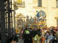 Imagen de una procesión religiosa en una ciudad histórica, con un carro de carnaval colorido y dorado frente a un edificio blanco. La gente observa la procesión, algunas con chalecos reflectantes.