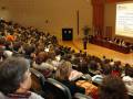 A una audiencia en un auditorio lleno, escuchando a un orador en el escenario. La pantalla grande proyecta una presentación con información sobre créditos ECTS y la Universidad de Salamanca. La audiencia está sentada en filas, con algunos estudiantes levantando sus teléfonos móviles. La sala está iluminada con una iluminación cálida y la decoración es simple, enfocándose en el contenido de la presentación.