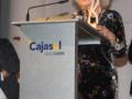 A una mujer con cabello blanco y largo, vestida con un traje floral, sosteniendo un trofeo mientras se encuentra en una plataforma de conferencia con el logotipo "Cajasol" visible.