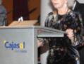 Una mujer con cabello blanco y largo, vestida con un traje floral negro, se encuentra en una conferencia o evento. Está frente a un podio con la marca "Cajasol" y el texto "BANCA CIVICA". La imagen parece ser de un evento formal, posiblemente una presentación o discurso.