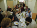Una larga fila de personas sentadas en mesas, compartiendo comida y conversando. La escena parece ser un comedor o evento social en una sala con paredes blancas y colores.