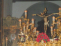 Imagen de una procesión religiosa con estatuas doradas y un crucificado en el centro, rodeado de flores rojas. La escena se desarrolla dentro de una iglesia con un techo de madera y una estructura de luces.