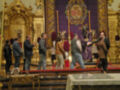 En una iglesia, un grupo de personas observa con atención a un sacerdote que sostiene una cruz. El altar está adornado con velas altas y un escudo heráldico en el fondo. La decoración dorada y la presencia de fieles sugieren una celebración religiosa importante.