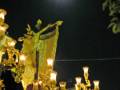 Estatua de Cristo en procesión nocturna, iluminada por una luna brillante y rodeada de árboles, con un cielo estrellado en el fondo.