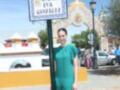 Una mujer en un vestido verde posa frente a una placa que indica la Avenida Eva González. En el fondo, se ve un arco con una bandera y varios vehículos estacionados.