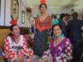 Una mujer en traje flamenco se levanta para tomar una copa mientras otras dos mujeres vestidas con trajes de flamenca se sientan en una mesa. La imagen muestra un ambiente festivo con decoración tradicional y personas sonrientes.