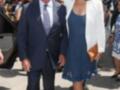 Una pareja camina juntos, el hombre lleva traje negro y corbata azul mientras la mujer usa vestido azul con blazer blanco. Ambos parecen estar en un evento formal al aire libre, posiblemente una celebración o reunión importante.