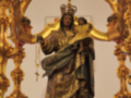 Estatua religiosa de una figura venerada, posiblemente una santa o virgen, con vestimenta ricamente ornamentada y un manto dorado. La imagen está rodeada por detalles dorados y espejos, sugiriendo un entorno de iglesia o templo.