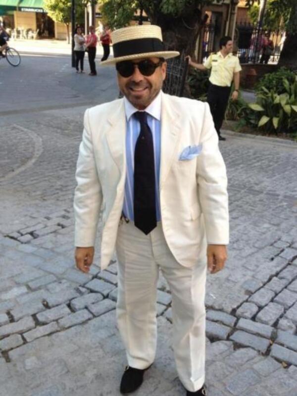 Un hombre vestido con un traje blanco y sombrero de paja camina por una calle adoquinada. Detalles: traje blanco, sombrero de paja, corbata negra, pantalones blancos.