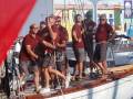 Grupos de personas vestidas con camisetas rojas y pantalones cortos, posiblemente de una misma asociación deportiva, están en un barco. Algunos llevan gafas de sol y otros tienen banderas ondeando en el fondo. El ambiente parece ser festivo, posiblemente una celebración o reunión de equipo.