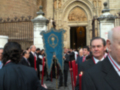 Una procesión religiosa en una catedral, con personas vestidas formalmente y un escudo de armas azul en el fondo.