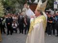 Un hombre vestido con un manto blanco y tocado con un sombrero de copa celebra una ceremonia religiosa en la calle. Detrás de él, hay personas vestidas con uniformes y otros individuos observando la celebración. La imagen muestra una escena festiva o religiosa en un entorno urbano con árboles y edificios al fondo.