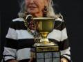 Una mujer con cabello blanco largo y una camiseta a rayas posa orgullosamente sosteniendo un trofeo de la competición "Rispresso".