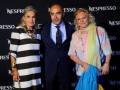 Tres personas posan frente a un fondo negro con el logo de Nespresso. La persona en medio viste una chaqueta negra y corbata, mientras que las dos personas a los lados llevan vestidos de colores vibrantes y accesorios.