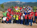 Una gran familia de personas, incluyendo niños y adultos, está posando para una foto en un entorno rural con montañas al fondo. Todos llevan camisetas rojas y amarillas, y algunos niños están vestidos con uniformes deportivos. La escena es soleada y la naturaleza parece ser el centro de atención en esta reunión familiar.