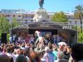 Una multitud congregada en un escenario al aire libre, con una estatua monumental en el centro y edificios de fondo. La gente está disfrutando de un evento público, con algunos levantando sus manos y otros mirando hacia el escenario. El cielo es claro y soleado, lo que añade un ambiente festivo a la escena.