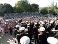 Un grupo de marineros en uniforme blanco y negro toca música en un escenario al aire libre. La audiencia, compuesta por personas de todas las edades, observa el concierto desde los asientos del estadio. La imagen captura una celebración festiva en un día soleado, con árboles y edificios al fondo.