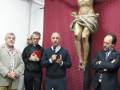En una iglesia, tres hombres observan con respeto a un Cristo crucificado en la pared roja. Un hombre sostiene una libreta, otro toca su corbata y el tercero mira hacia abajo. En la mesa, hay un cáliz y flores blancas.