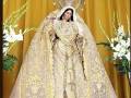 Imagen de una Virgen María vestida con un manto blanco y dorado, rodeada por flores y una cortina amarilla.