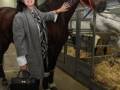 Una mujer con abrigo gris y accesorios de moda posa junto a un caballo en una feria o exposición. La imagen muestra detalles de la moda y el ambiente de la feria, destacando la interacción entre la mujer y el animal.