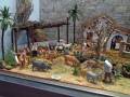 Un pequeño escenario de animales en miniatura, con una figura humana en el centro. Hay varios animales como ovejas y cerdos, junto con una figura humana en el centro. La escena está rodeada de vegetación y hay un edificio en la parte superior derecha.