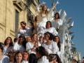 Una procesión religiosa con una imagen de la Virgen en el centro, rodeada por jóvenes vestidas de blanco y cabellos dorados. El cielo azul claro brilla sobre la escena, enfatizando el ambiente festivo y religioso.
