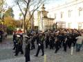 Fotografía de una procesión militar en un entorno urbano con uniformes militares, bandas y gente observando.