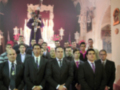 Un grupo de hombres vestidos con trajes formales se encuentran en una iglesia, posiblemente durante un evento religioso o ceremonial. La decoración interior incluye candelabros y una cruz visible en el fondo.