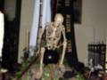 Un esqueleto sentado en un altar de Halloween, rodeado por calabazas y velas encendidas. En el fondo, una pintura de un fantasma y un cartel que dice "Supernatural".
