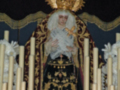 Imagen de un personaje vestido con trajes ceremoniales, posiblemente religiosos, sentado en un trono. El personaje lleva una corona y tiene cabello blanco largo. En el fondo hay velas encendidas que iluminan la escena. La imagen parece ser de una celebración o procesión religiosa.