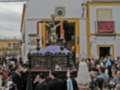 Imagen de una procesión religiosa con un crucifijo en el centro, rodeado por fieles y oficiales. El edificio al fondo es blanco con detalles dorados, y hay banderas y decoraciones en el exterior.