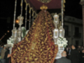 Imagen de una procesión religiosa nocturna con una imagen venerada envuelta en un manto dorado y bordado, acompañada por personas vestidas de negro.