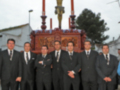 Un grupo de hombres en trajes formales posa frente a una imagen religiosa, probablemente durante una procesión o ceremonia.