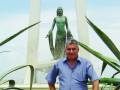 Un hombre posa frente a una estatua de una mujer en un monumento, rodeado por plantas y con un cielo azul claro de fondo.