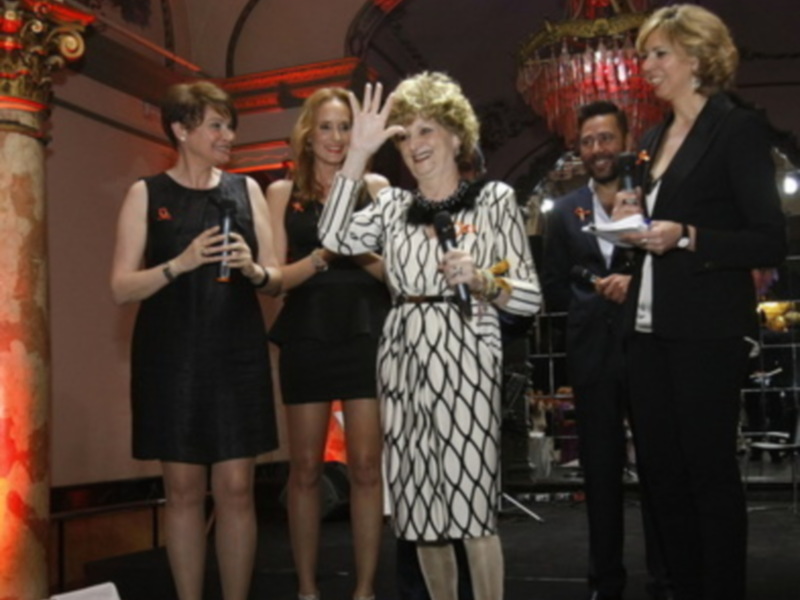 Una mujer con una camisa estampada y un vestido negro, junto a otras personas en un evento formal. La escena tiene una atmósfera festiva con luces y decoración.