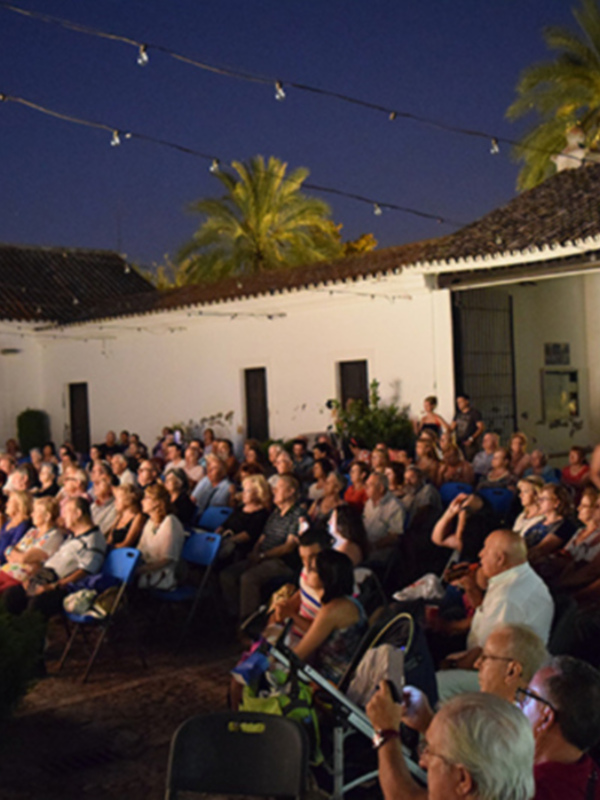 Una multitud de personas asiste a un evento nocturno al aire libre en un entorno arquitectónico colonial, con palmeras y una estructura con tejas rojas en el fondo.
