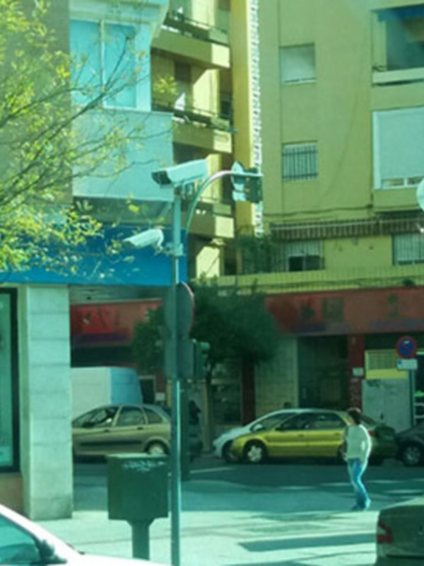 Vista de una calle urbana con edificios, árboles y vehículos en movimiento.