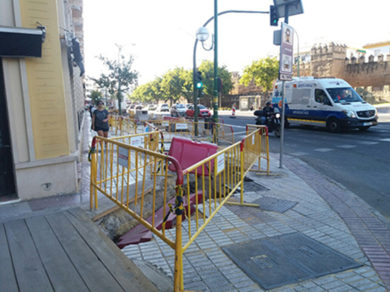 Trabajos de reparación en el suelo de la calle, con barreras amarillas y una ambulancia en el fondo.