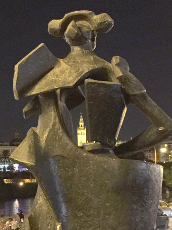 Escultura de bronce en la noche, con un fondo nocturno que incluye una torre iluminada y árboles verdes. La escultura parece ser una figura femenina con un sombrero y una prenda de vestir. El ambiente es谧谧,暗示着一种宁静和优雅。