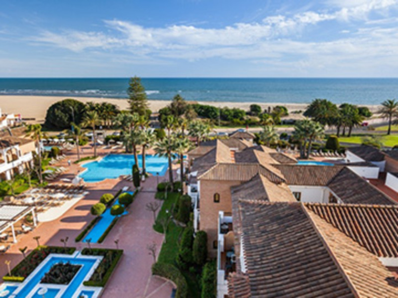 Vista panorámica de un resort con piscina, palmeras y vistas al mar bajo un cielo azul claro.